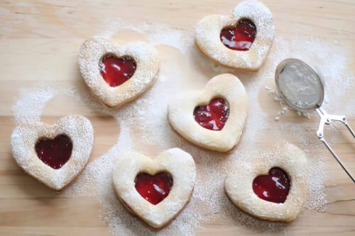 Linzer cookies, heart-shaped and filled with red jam, are artfully arranged on a wooden surface, dusted with powdered sugar. A mesh strainer lightly coated in sugar rests nearby.