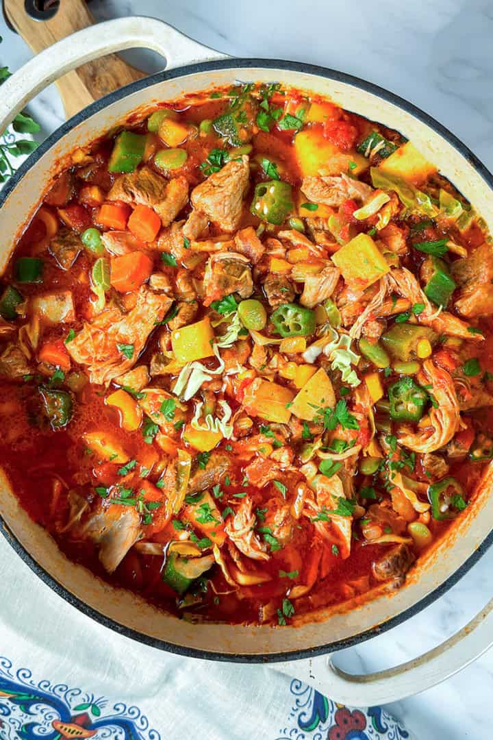 A pot of hearty burgoo stew with carrots, potatoes, okra, zucchini, and fresh herbs simmering in a tomato-based broth on a white surface.