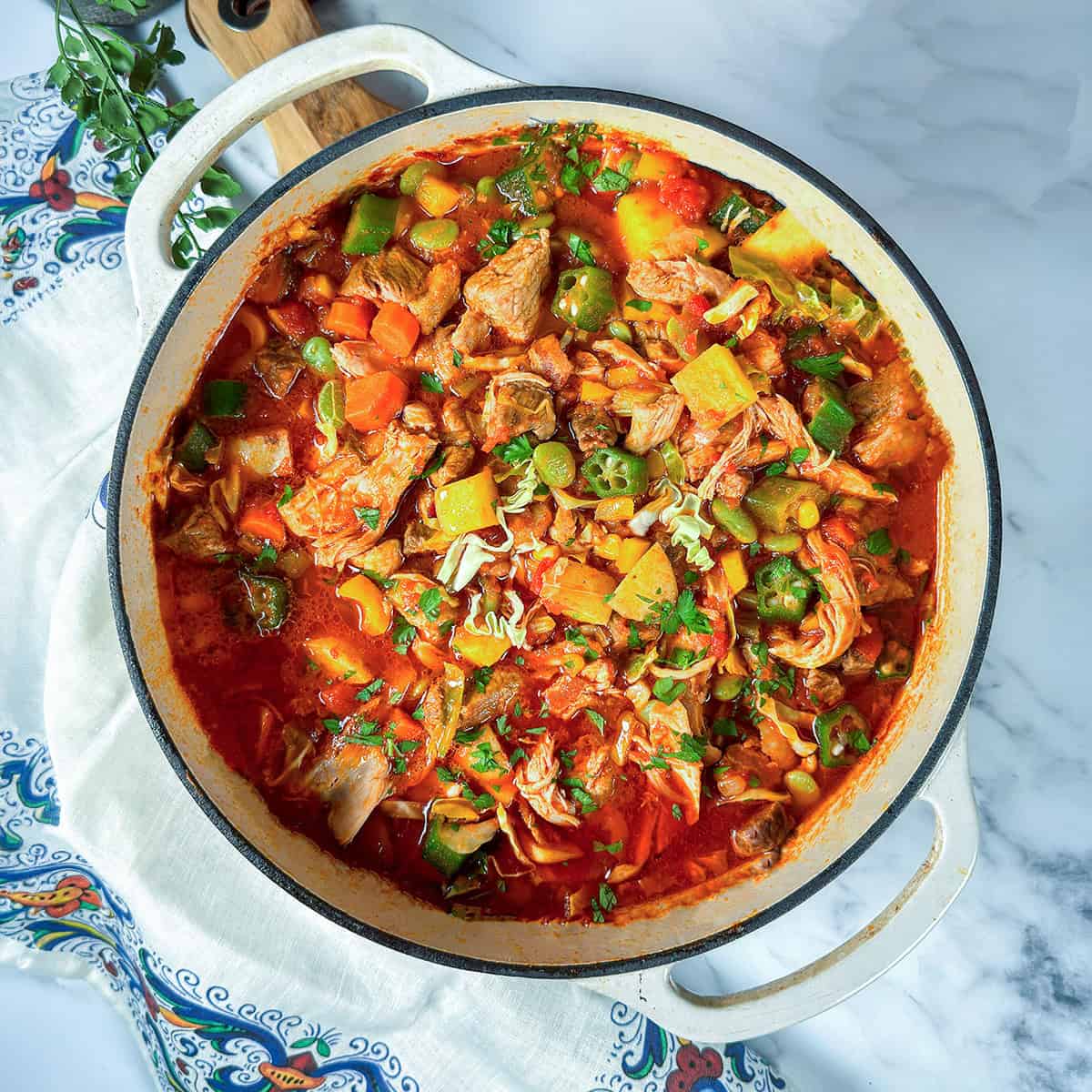 A pot of hearty burgoo stew with carrots, potatoes, okra, zucchini, and fresh herbs simmering in a tomato-based broth on a white surface.