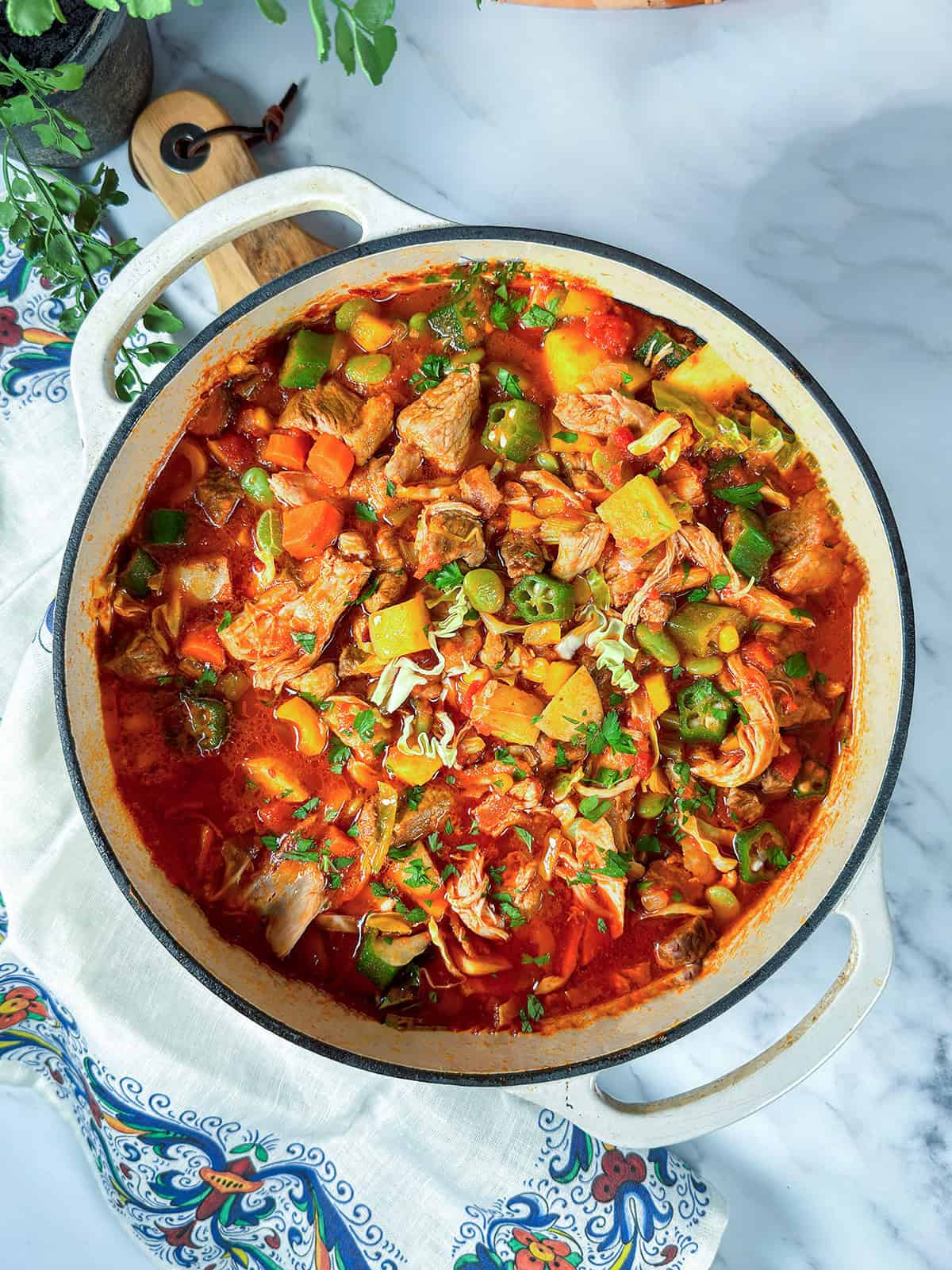 A pot of burgoo stew with chunks of meat, vegetables, and herbs in a tomato-based broth sits invitingly on a white cloth with a patterned edge.