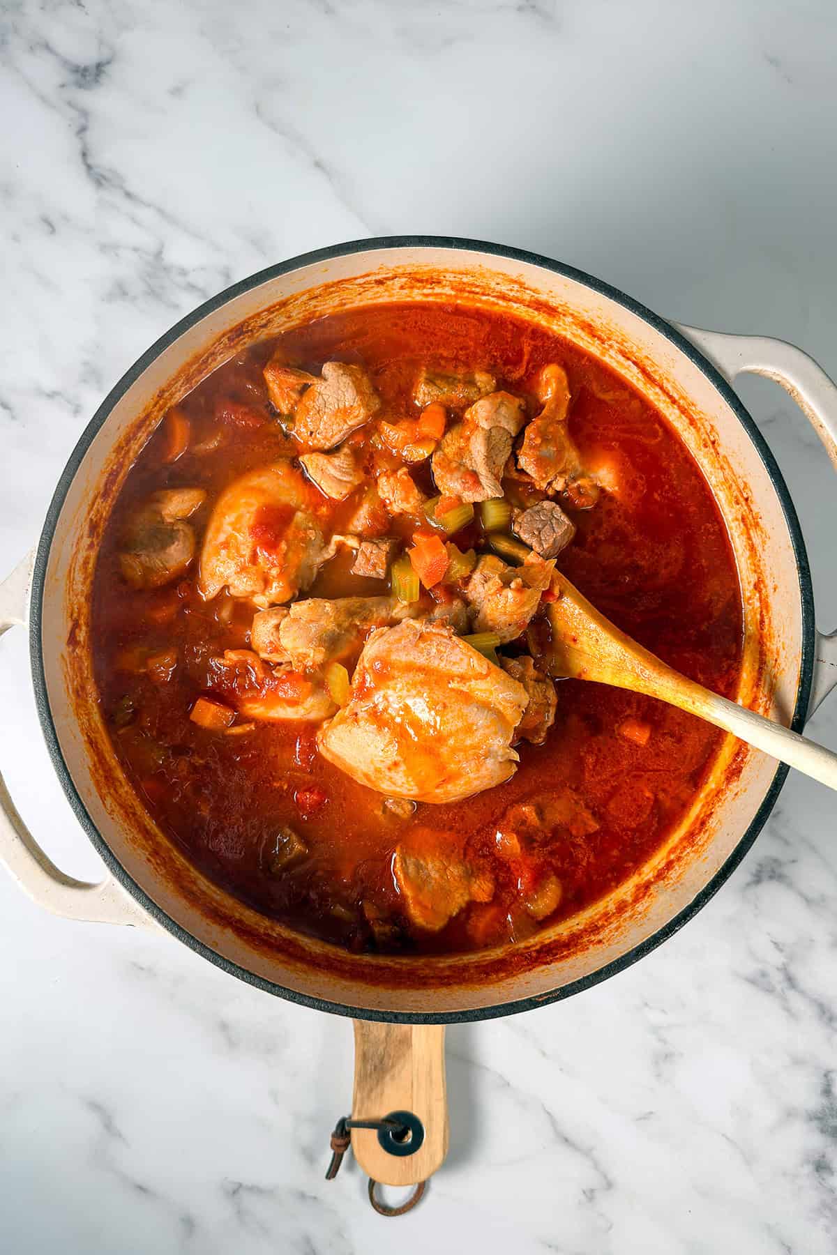 A pot filled with a hearty Burgoo, a tomato-based stew brimming with chunks of meat and vegetables with broth and seasoning being added. 