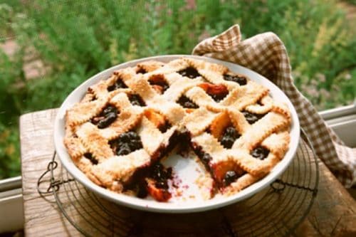 A blueberry peach pie with a lattice crust sits on a cooling rack by a window, one slice missing. A checked cloth is draped beside the dish.