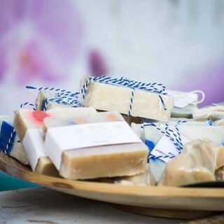 A wooden tray holds several bars of handmade oatmeal soap, each wrapped with paper and tied with blue and white string. Other small pieces from the oatmeal soap recipe are also visible on the tray.