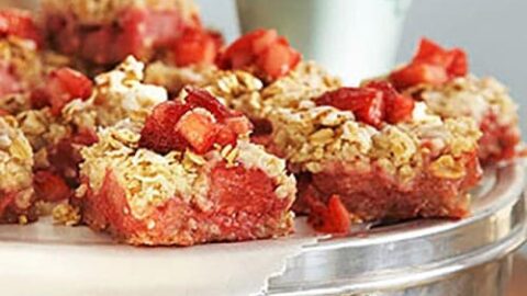 Close-up of Strawberry Rhubarb Bars on a tray. The bars have a chunky, crumbly texture with visible pieces of strawberries, rhubarb, and oats, creating a layered appearance. The background includes a blurred hint of a white and light green object, possibly a cup or similar item.