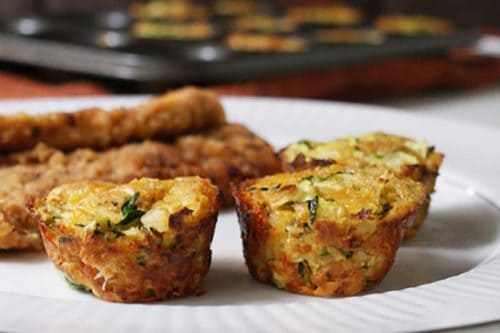 Three baked zucchini muffins, reminiscent of savory zucchini tots, served on a white plate with breaded meat and a tray of more muffins in the background.