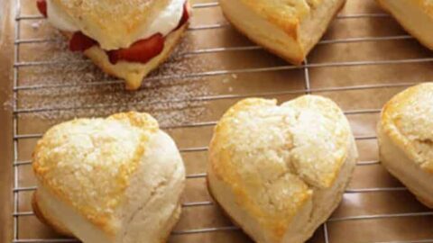 Valentine strawberry shortcake biscuits rest on a cooling rack, heart-shaped and filled with cream and strawberries, topped with cherries. The tray sits on a rustic wooden surface.