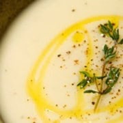 A close-up of a creamy potato soup garnished with a swirl of olive oil, fresh thyme, and a sprinkle of black pepper. The soup is served in a rustic brown bowl.