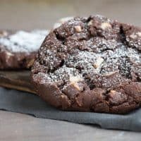 Two large Double Chocolate Chip Cookies with white chocolate chips and a dusting of powdered sugar are placed on a wooden board over a dark cloth.