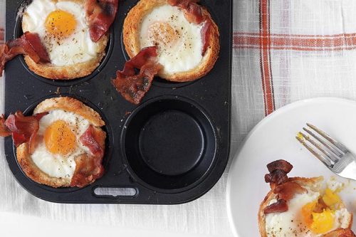 A muffin tin holds three egg and bacon cups with toast; one cup is missing, partially eaten on a white plate beside a fork.