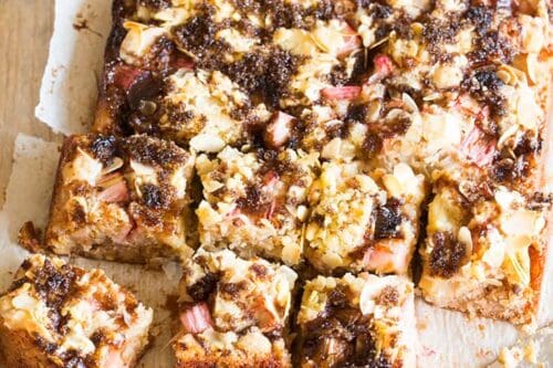 A square Rhubarb Cake topped with crumble and almonds is cut into pieces on parchment paper. A cake server with crumbs rests nearby, and a small bowl of sliced almonds sits in the background.