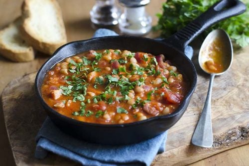 A cast-iron skillet filled with Skillet Baked Beans and tomato stew, garnished with chopped herbs, sits on a cloth with bread slices, a spoon, and salt and pepper shakers nearby.