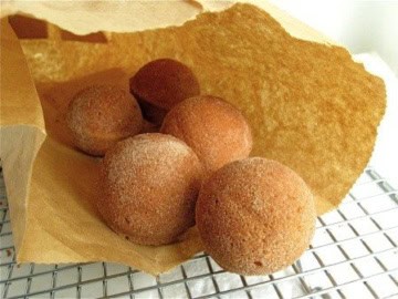 Five round, brown baked doughnut holes coated in sugar are partially visible inside a torn brown paper bag, resting on a metal cooling rack.