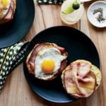 A black plate with an open-faced sandwich, topped with savory ham, a sunny-side-up egg, and melted cheddar cheese, sits on a wooden table next to a drink and a small bowl of pepper.