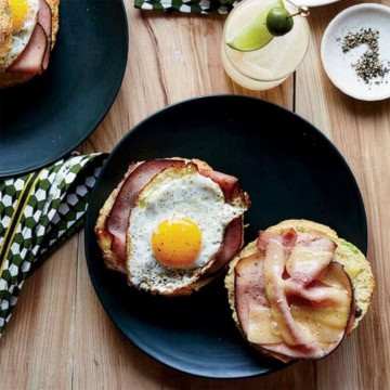A black plate with an open-faced sandwich, topped with savory ham, a sunny-side-up egg, and melted cheddar cheese, sits on a wooden table next to a drink and a small bowl of pepper.