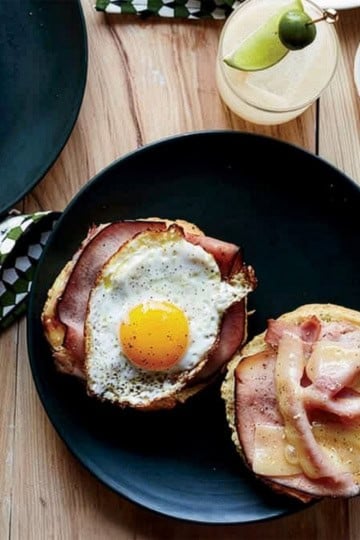 A black plate with an open-faced sandwich, topped with savory ham, a sunny-side-up egg, and melted cheddar cheese, sits on a wooden table next to a drink and a small bowl of pepper.