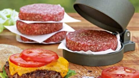 Raw beef burger patties and a burger press sit beside a finished burger with lettuce, cheese, and tomato on a wooden board—perfect for summer barbecues or as inspiration for Father's Day gift ideas. Sliced tomatoes and lettuce are also visible.