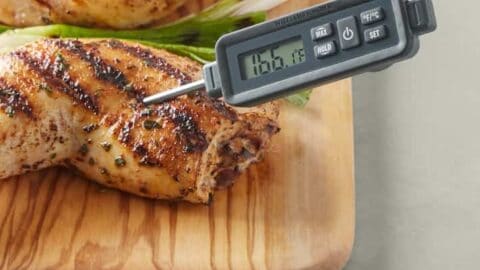 A digital meat thermometer, a perfect addition to father's day gift ideas, is inserted into a grilled chicken leg on a wooden board, displaying the temperature. Green onions and another piece of grilled chicken are in the background.