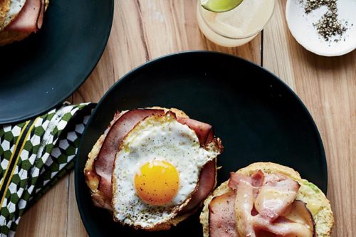 Two black plates with breakfast sandwiches: one with ham and a sunny-side-up egg, and the other with ham and melted cheese. A folded napkin, a drink with a lime and olive garnish, and a small dish of salt and pepper are also on the wooden table.