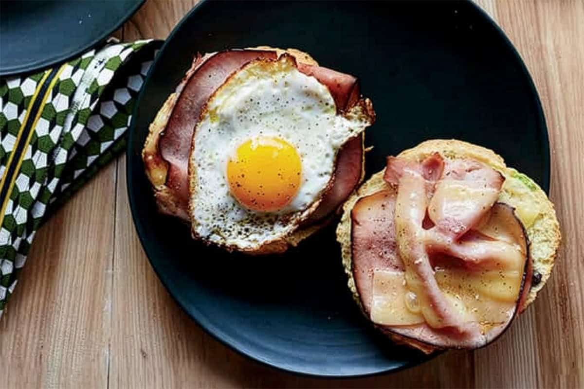 Two halves of ham biscuit sandwiches on a black plate-one with ham and a fried egg, the other with ham and melted cheese-served on a wooden table.