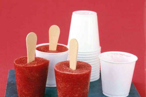 Three red popsicles with wooden sticks in white paper cups are placed on a blue stone slab against a red background. Nearby, there is a stack of white paper cups and one empty white cup-ideal for trying out your favorite summer popsicle recipes.