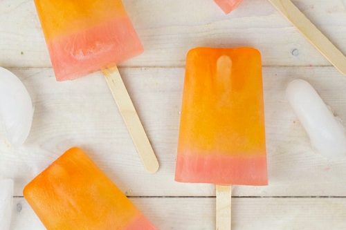 Four orange and pink gradient popsicles are scattered on a light wooden surface, surrounded by ice cubes. Text at the bottom right reads "Easy Orange Pink Lemonade Popsicles" and a watermark at the top left reads "formulamom.com." Perfect for summer popsicle recipes!