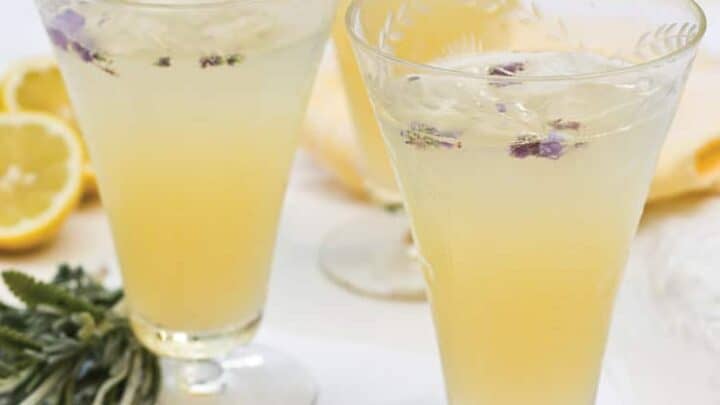 Three elegant glasses filled with a light yellow homemade lemonade are garnished with lavender flowers. Slices of lemon and sprigs of lavender are scattered around the base of the glasses on a white surface. The overall scene is bright and refreshing.