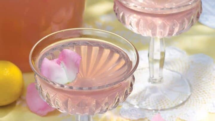 Two elegant glasses of homemade pink lemonade with rose petals are placed on a lace doily. A pitcher of the same refreshing homemade lemonade, adorned with floating rose petals, is in the background. Fresh lemons and lemon slices on a dish are also visible, adding a bright, summery feel.