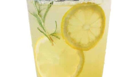 A clear glass filled with homemade lemonade is garnished with two lemon slices and a sprig of rosemary. The drink has a refreshing, pale yellow hue and is set against a plain white background.