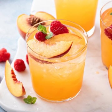 A glass of raspberry peach iced tea garnished with peach slices, raspberries, and mint leaves sits on a white surface with additional fruit pieces nearby.