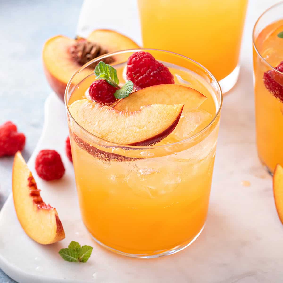A glass of raspberry peach iced tea garnished with peach slices, raspberries, and mint leaves sits on a white surface with additional fruit pieces nearby.
