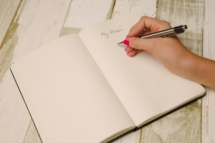 A person writes "My plan:" with a pen on a grid notebook open to a blank page, resting on a light wooden surface—perfect for organized people to track daily habits and encourage easy organization.