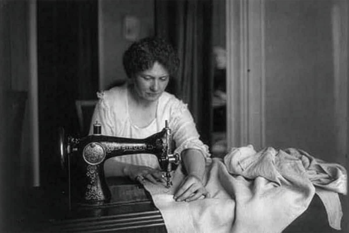 A woman sits at a vintage sewing machine, guiding a flour sack under the needle in a softly lit room.
