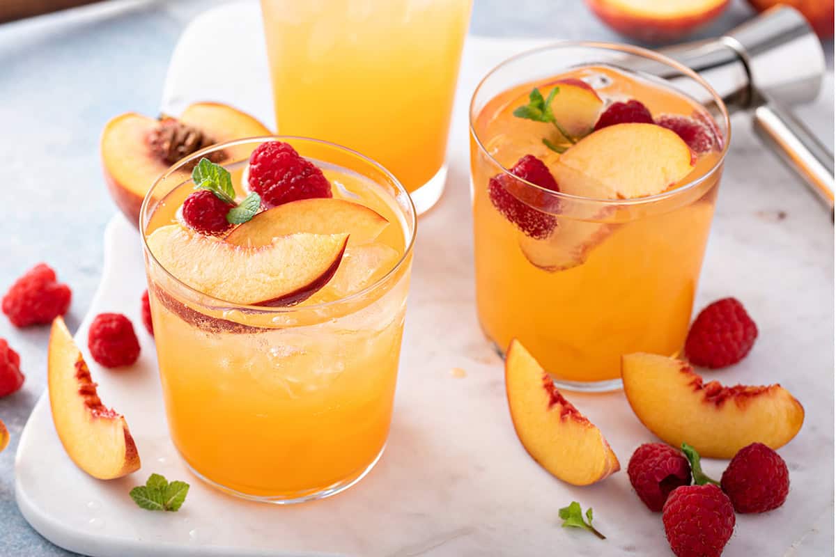 Two glasses of raspberry peach iced tea, garnished with fresh peach slices, raspberries, and mint, rest on a marble surface.