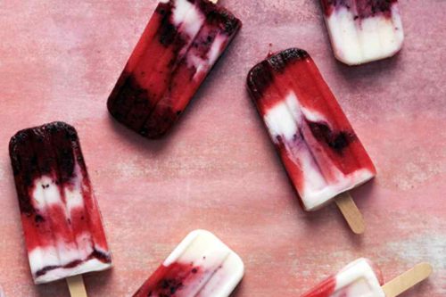 A photograph showing six summer popsicles scattered on a pink surface. The popsicles are distinctively red, white, and purple, boasting a marbled effect from blending the different colors. Each has a wooden stick handle, perfect examples of delightful summer popsicle recipes.
