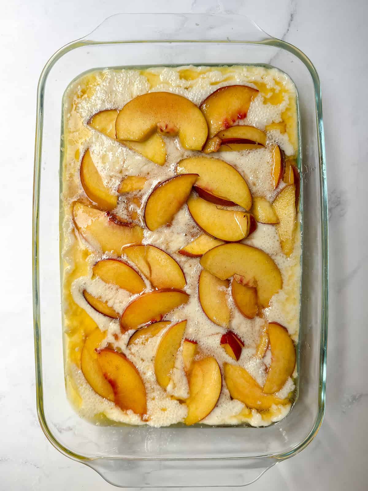 Sweetened peaches spooned over the butter and cobbler batter in a baking dish.