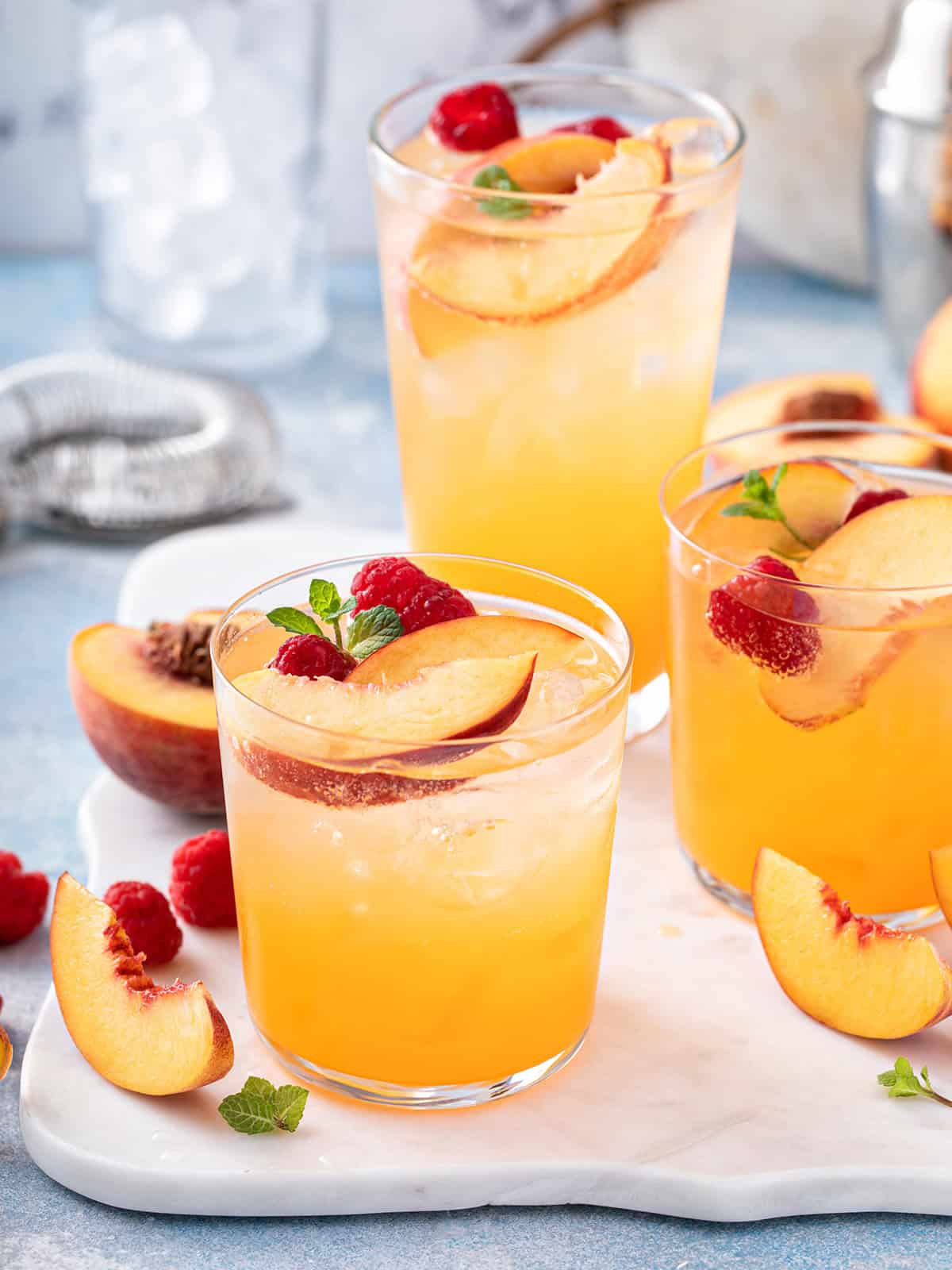 Three glasses of raspberry peach iced tea garnished with fruit slices and mint on a white surface.