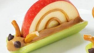 A fall healthy snack depicting a snail made from a red apple slice for the shell, peanut butter as the body, a green celery stick as the base, pretzel sticks for antennae, and chocolate chips for eyes, all creatively arranged on a plate.