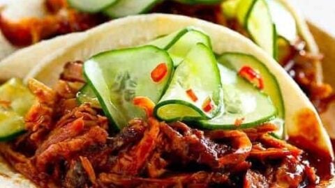 Two soft tortillas filled with shredded, seasoned meat and topped with sliced cucumbers, red chili flakes, and a drizzle of sauce, placed on a wooden plate. These tacos follow authentic street food recipes, looking fresh and appetizing with their mix of textures and vibrant colors.