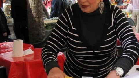 A woman wearing a black hat and striped shirt serves slices of large, round flatbread from a metal tray at an outdoor market. Red and white striped tents and other people are visible in the background, creating a vibrant scene perfect for exploring street food recipes.