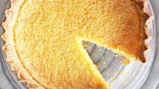 An award-winning lemon chess pie sits in a glass dish with one slice missing, viewed from above on a gray, circular patterned surface.