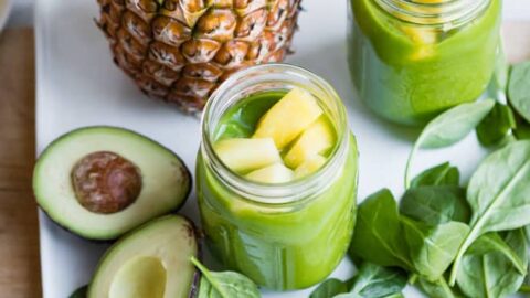 A pineapple, sliced avocado, spinach leaves, and two mason jars of weight loss smoothies topped with pineapple pieces are arranged on a white tray.