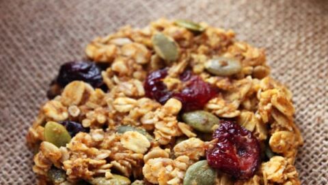 A close-up of a homemade oatmeal cookie on a burlap surface. The cookie is packed with various seeds, nuts, and dried fruits, including pumpkin seeds and cranberries, giving it a textured and wholesome appearance. Perfect for fall healthy snacks, other similar cookies are partially visible in the background.