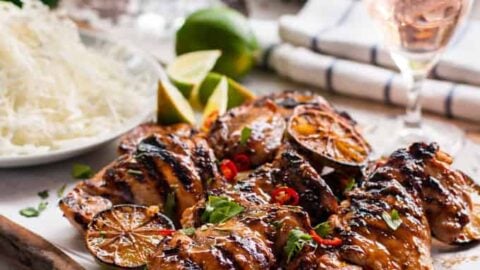 Grilled chicken pieces garnished with lime slices, chopped red chili, and fresh herbs, served on a wooden board. Nearby, there's a bowl of rice, a fork, knife, and a small dish of chopped herbs, accompanied by glasses of beverages. The relaxed outdoor dining setting captures the essence of street food recipes.
