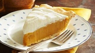 A slice of award-winning pie with a creamy topping sits on a decorative plate, accompanied by a fork and a cup of coffee on a brown table.