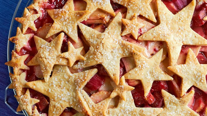 An award-winning fruit pie with a golden crust, topped with various sizes of star-shaped cutouts, sits on a wire cooling rack over a blue surface.