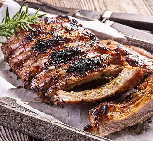 Grilled Baby Back Ribs with a shiny glaze are arranged on parchment paper, garnished with a sprig of rosemary, and served on a rustic tray with a knife and fork in the background.