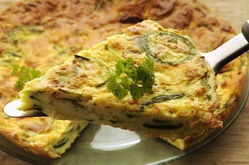 A slice of zucchini frittata with tomato and bacon rests on a serving spatula, garnished with parsley, held above the rest of the quiche in a glass dish.