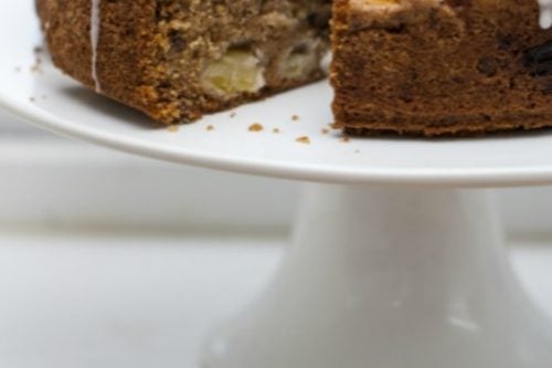 A round apple cake topped with glaze and chopped nuts sits beautifully on a white cake stand. A slice has been cut out, revealing the moist interior with visible apple pieces. The image text reads, "Autumn Essential Raw Apple Cake" and proudly features "31Daily.com" at the bottom.