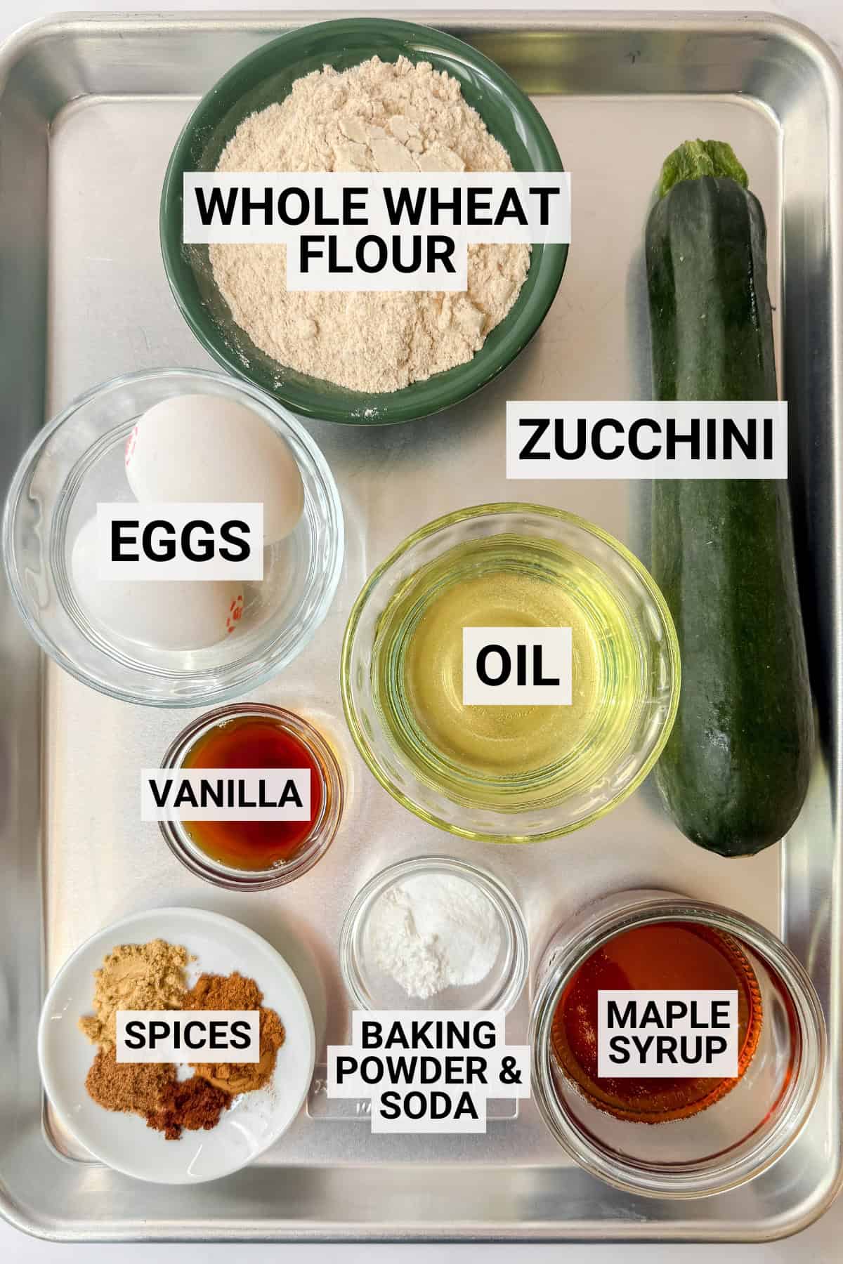 Baking tray with whole wheat flour, zucchini, egg, oil, vanilla, spices, baking powder and soda, and maple syrup in separate bowls-everything you need to prepare delicious muffins.
