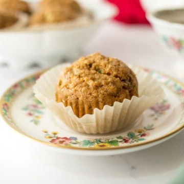A single muffin in a paper liner sits on a floral-patterned plate, with more muffins and a teacup blurred in the background, creating a cozy scene perfect for enjoying freshly baked muffins.